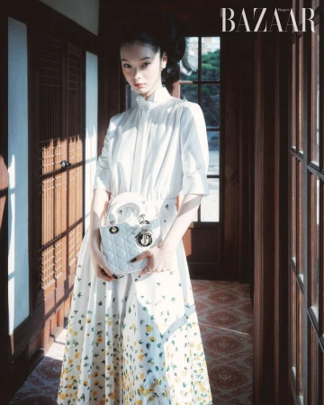 Elegant woman in a white floral dress holding a white purse, posing indoors with natural light coming through the window.