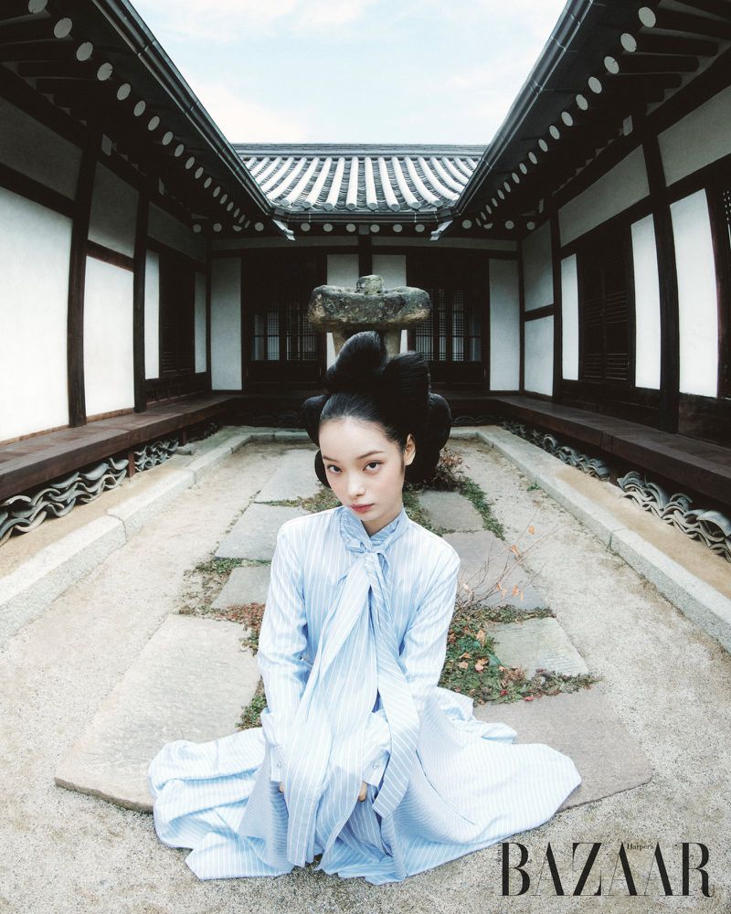 Fashion editorial shot of an Asian woman kneeling in a traditional Korean courtyard wearing a light blue striped dress with her hair styled up.