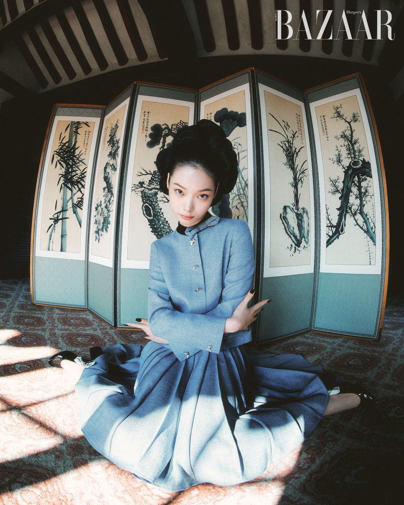 A woman sits on a patterned floor in front of a folding screen wearing a light blue outfit in a fashion editorial for Bazaar magazine.