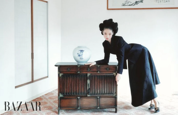 A woman in a black dress and elaborate hairstyle poses next to a wooden cabinet with a ceramic vase in a light-filled interior.