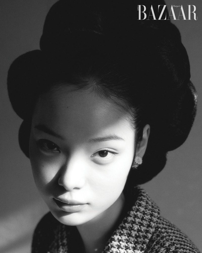 Black and white portrait of a young woman wearing a patterned jacket and an elaborate updo hairstyle, against a neutral background.