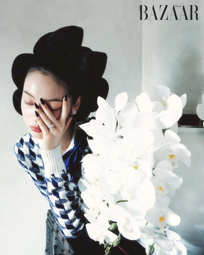East Asian woman in a sculptural black headpiece and patterned knit sweater posing beside white orchids with hand over face.