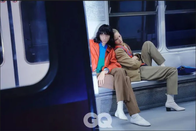 Two fashion models pose seated in a subway car, showcasing stylish blazers, slacks, and shoes in a photograph with cool, moody lighting.
