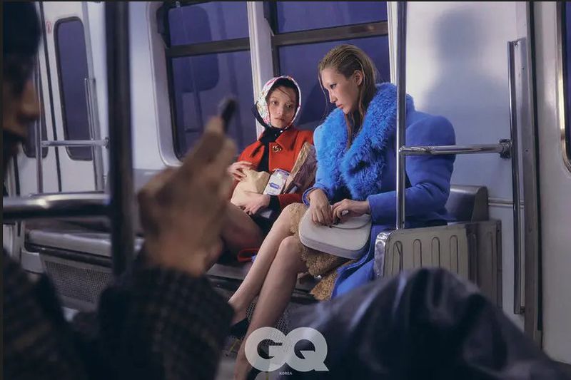 Two stylish women sit on a train wearing fashionable overcoats; one is red with a scarf and the other is blue with a fur collar.