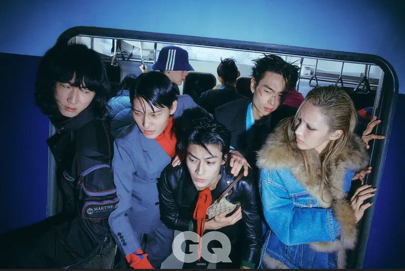 Fashion editorial shot of a diverse group of young adults on a train, showcasing trendy clothing and edgy makeup with moody lighting and GQ Korea logo.