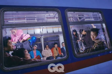 Models in vibrant high-fashion outerwear seated and standing inside a blue subway car, seen through train windows.