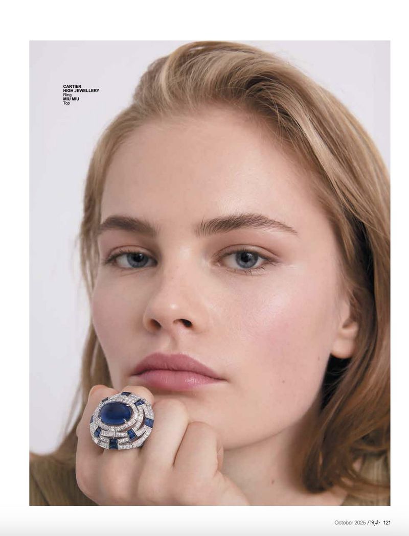 Close-up fashion shot of a blonde woman wearing a large sapphire and diamond ring, posing with her hand near her face in a studio.