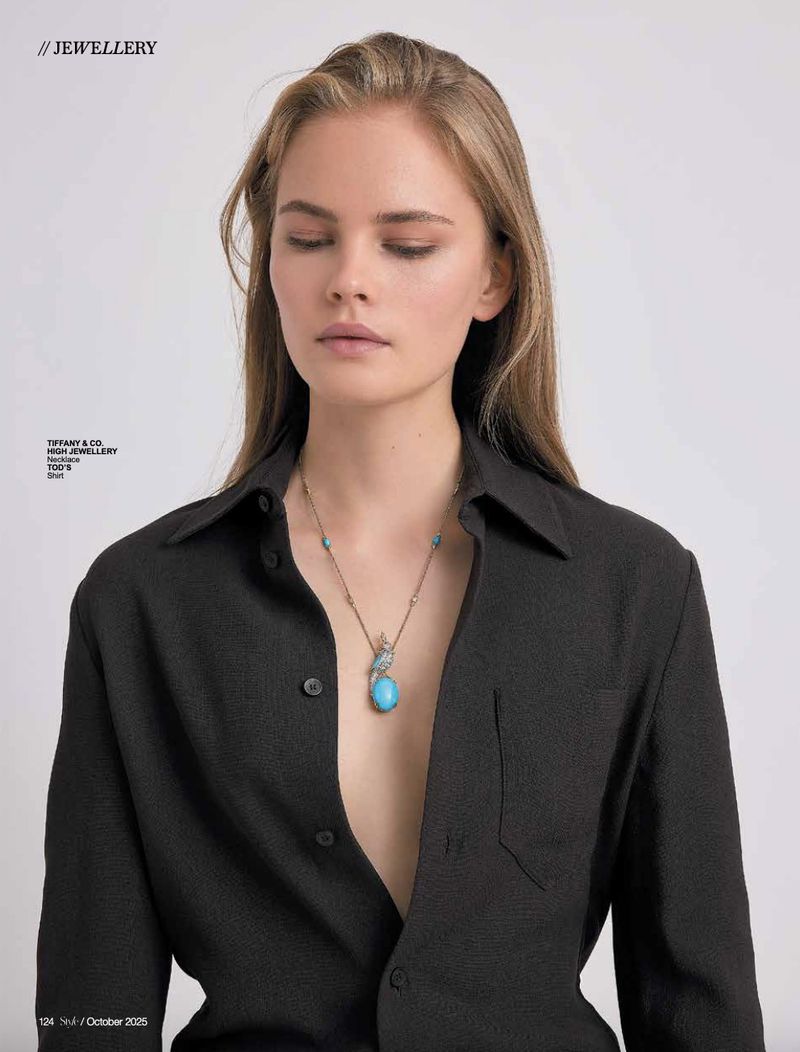 A woman with light brown hair wears a black open button-down shirt and a necklace with a large turquoise pendant against a white background.