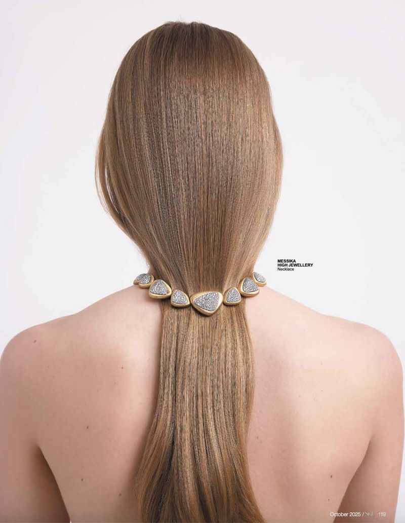 Back view of a woman with long brown hair wearing a triangle-shaped gold and diamond necklace against a clean white background.