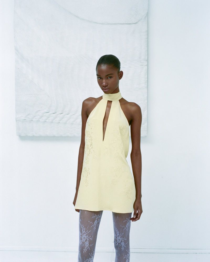 Elegant Black woman in a yellow halter minidress with lace tights, posing in a bright white studio with minimal decor. Fashion editorial.