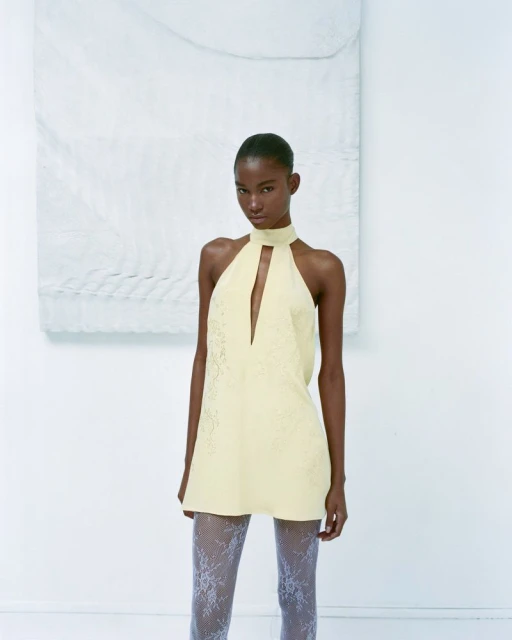 Elegant Black woman in a yellow halter minidress with lace tights, posing in a bright white studio with minimal decor. Fashion editorial.