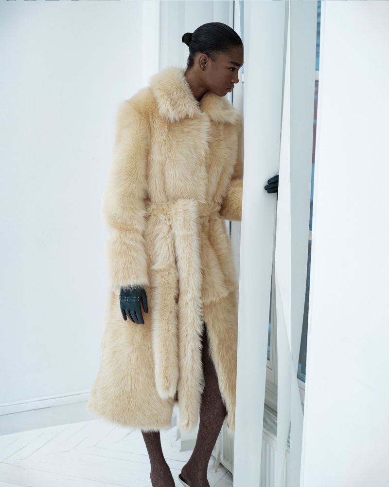 A lone woman in a fur coat and gloves poses by a set of white blinds against a white wall, captured in a studio fashion shot.