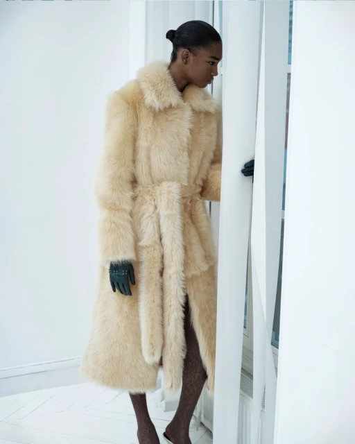A lone woman in a fur coat and gloves poses by a set of white blinds against a white wall, captured in a studio fashion shot.