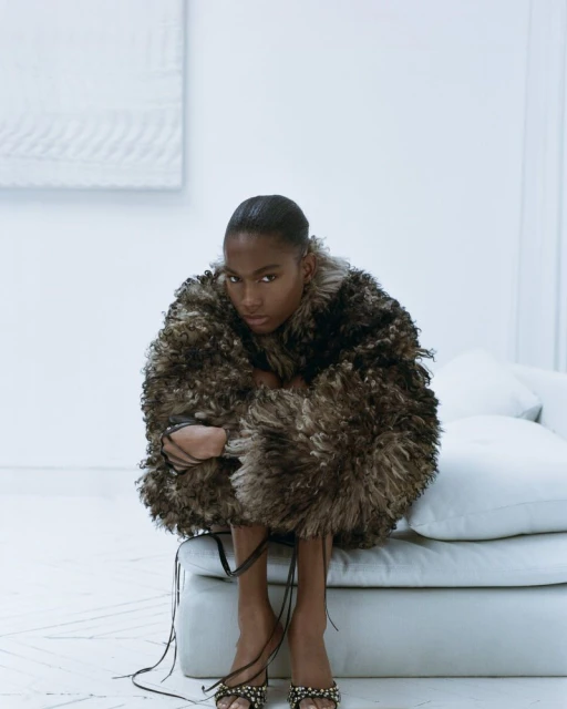 Dark-skinned woman in an elaborate brown feathered coat and beaded high-heeled sandals, sits on a white sofa against a minimalist white background.