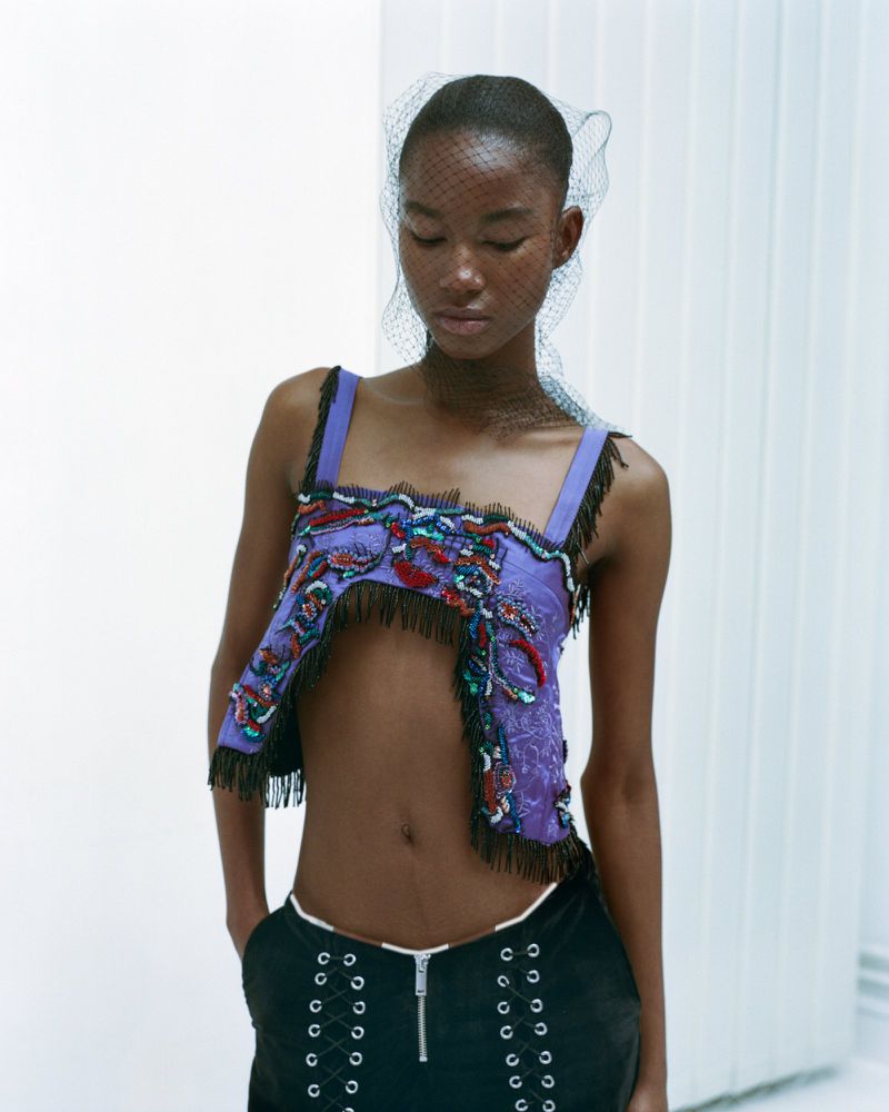 Fashion shot of a Black woman wearing an open-midriff top, black bottoms, and a net veil posing against a white background.