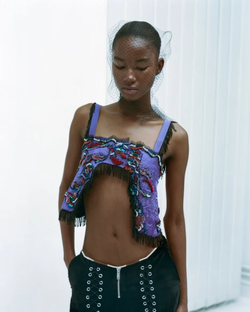 Fashion shot of a Black woman wearing an open-midriff top, black bottoms, and a net veil posing against a white background.
