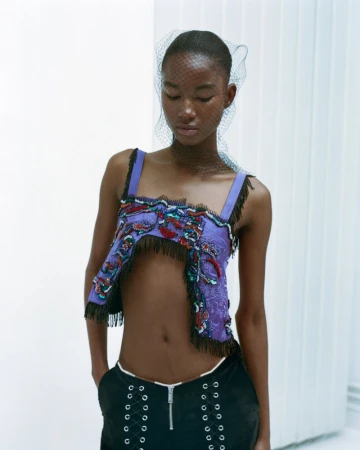 Fashion shot of a Black woman wearing an open-midriff top, black bottoms, and a net veil posing against a white background.