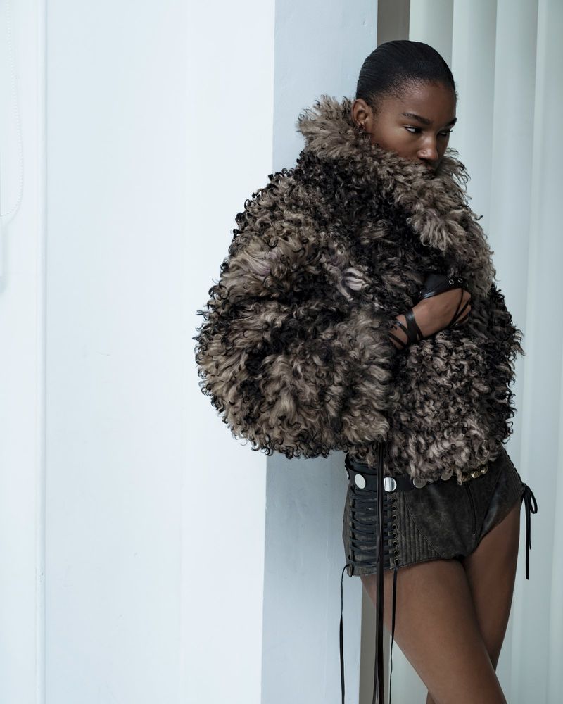 Fashion shot of a black female model in a sheepskin jacket and dark shorts posing against a bright wall in a studio setting.