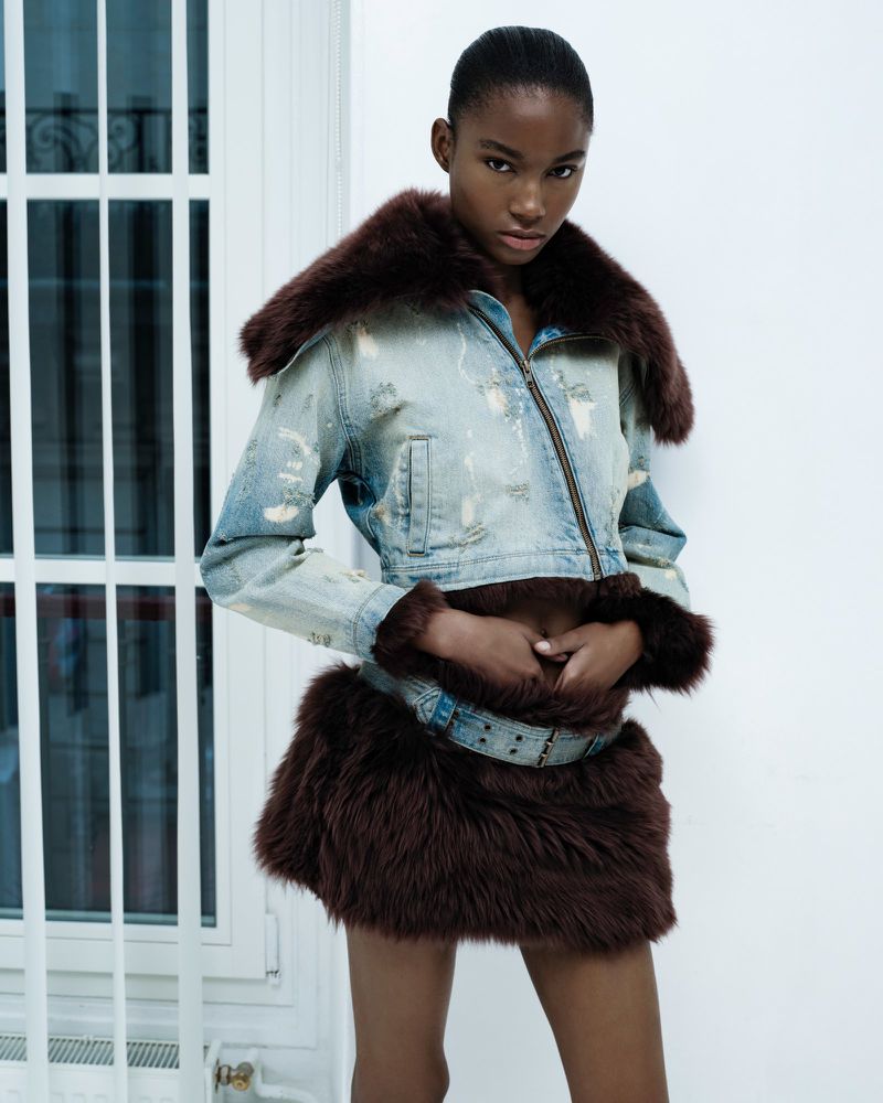 Woman posing in distressed denim jacket with fur trim and matching mini skirt against a white wall and window frame.
