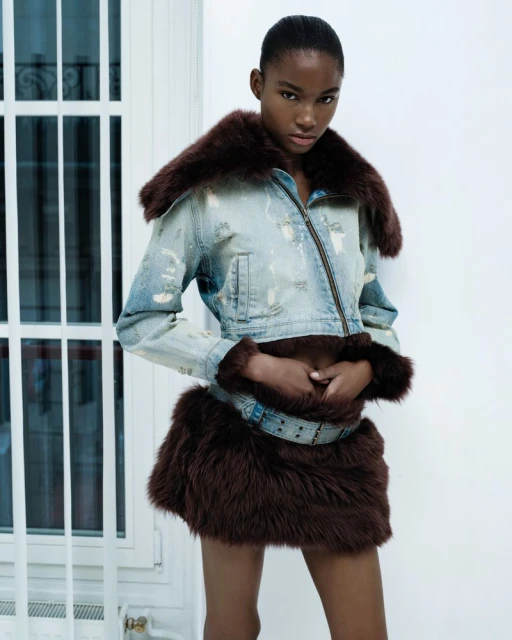 Woman posing in distressed denim jacket with fur trim and matching mini skirt against a white wall and window frame.