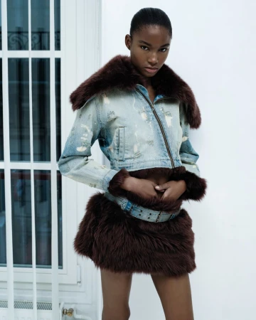 Woman posing in distressed denim jacket with fur trim and matching mini skirt against a white wall and window frame.