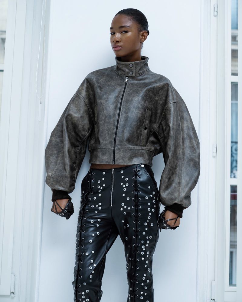 Woman in grey leather jacket and black leather pants with silver grommets posing against white wall in studio for fashion editorial shot.