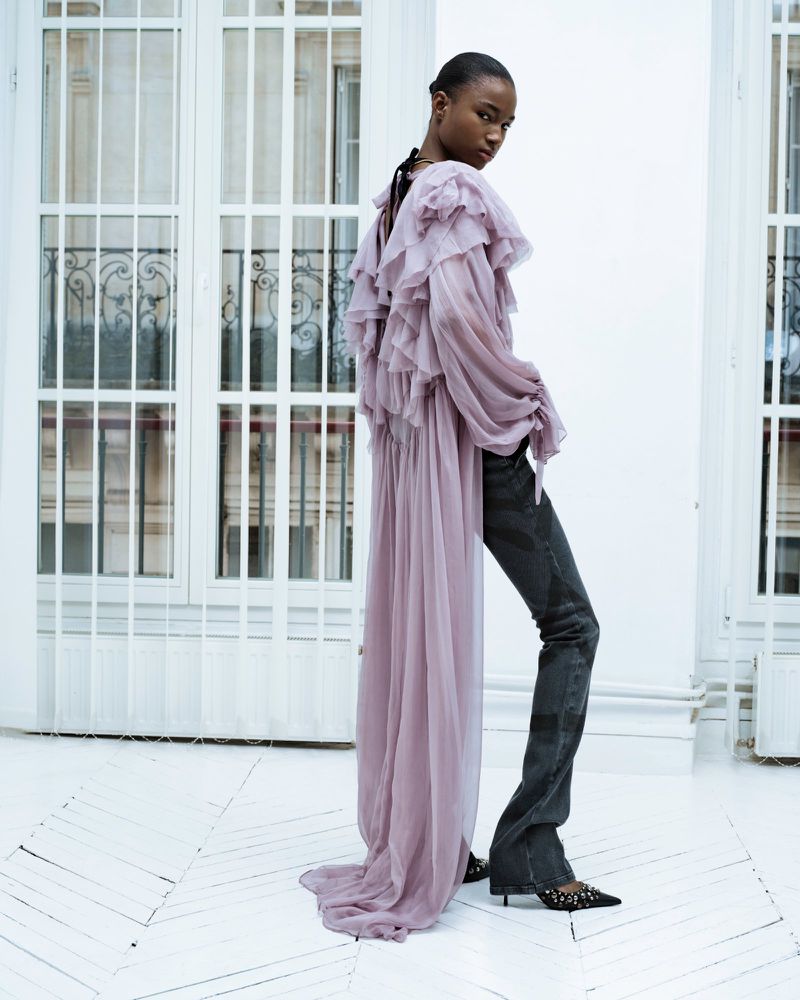 Fashion shot of a black woman with dark hair posing in a lavender ruffled top and dark denim pants in a white indoor space.