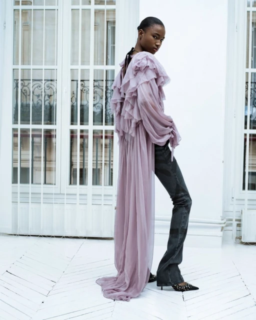Fashion shot of a black woman with dark hair posing in a lavender ruffled top and dark denim pants in a white indoor space.