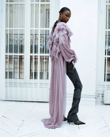 Fashion shot of a black woman with dark hair posing in a lavender ruffled top and dark denim pants in a white indoor space.