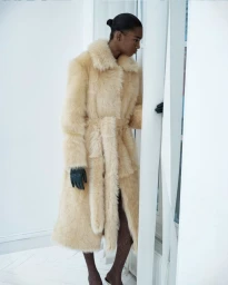 Model in a cream faux fur coat wearing dark gloves standing against a bright white background