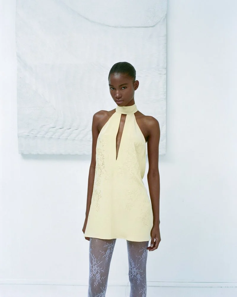 Model in a pale yellow halter dress and patterned tights standing against a white studio background for Maison Sel Lookbook