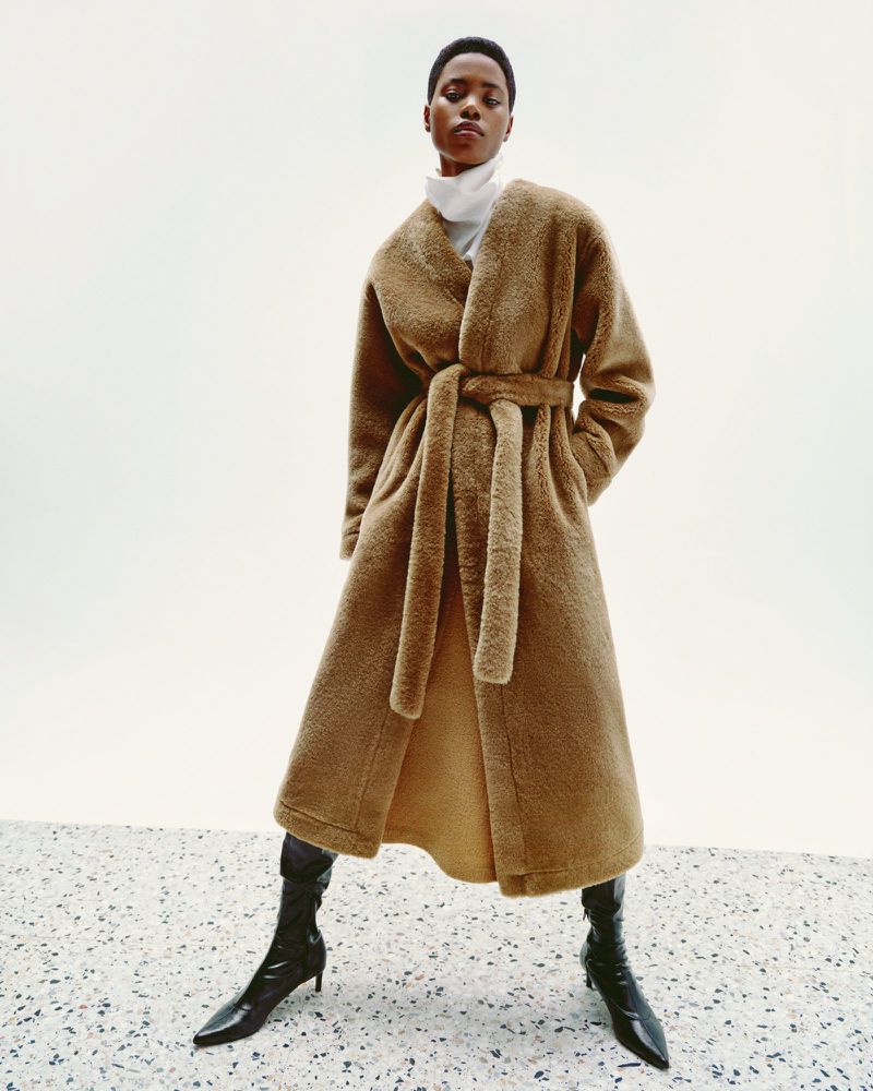 Elegant African-American woman in a plush brown wrap coat, black heeled boots poses against a white backdrop, fashion forward.