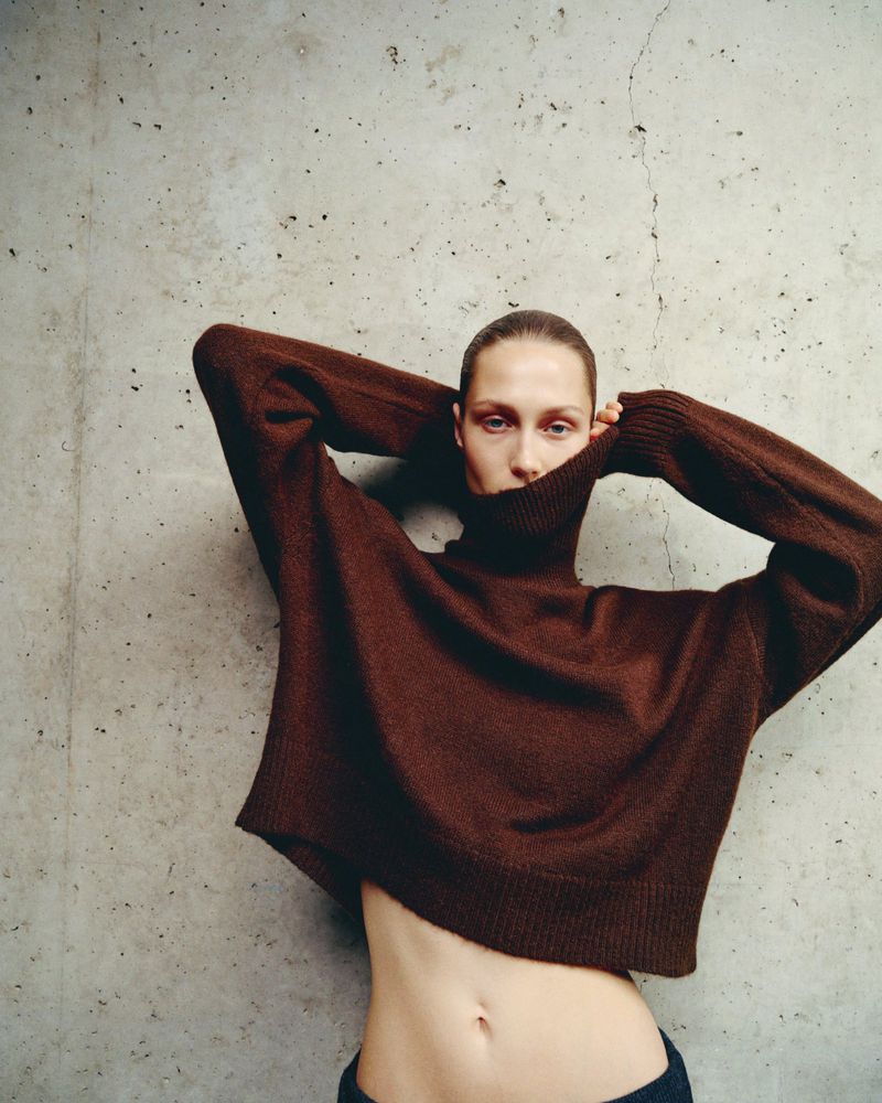 Fashion shot of a woman wearing a brown sweater against a concrete wall, hands behind her head, looking at the camera.