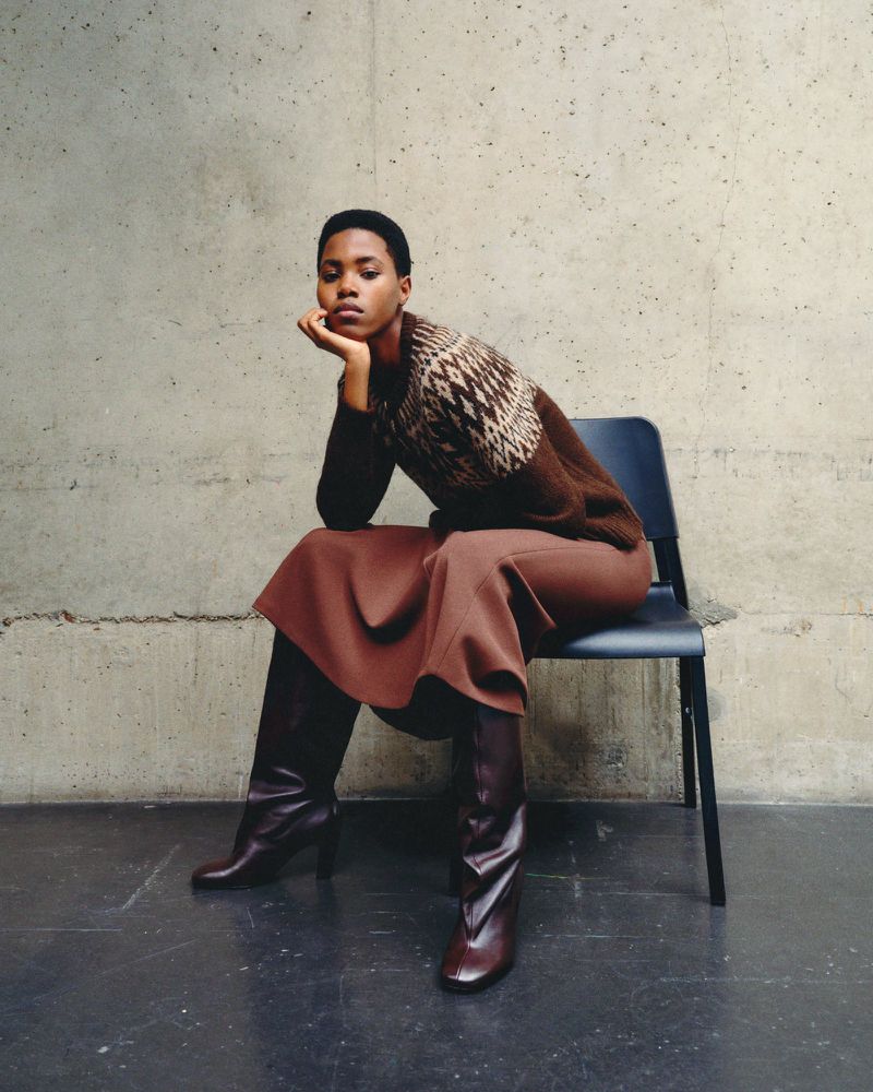 Model seated on a black chair wearing a brown patterned sweater, skirt, and dark boots against a concrete wall