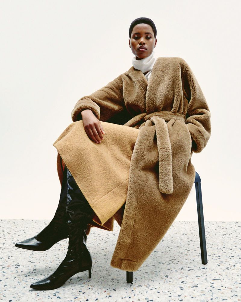 An adult woman with short dark hair sits on a stool in a tan coat and black boots against a white background, posing for a fashion shot.