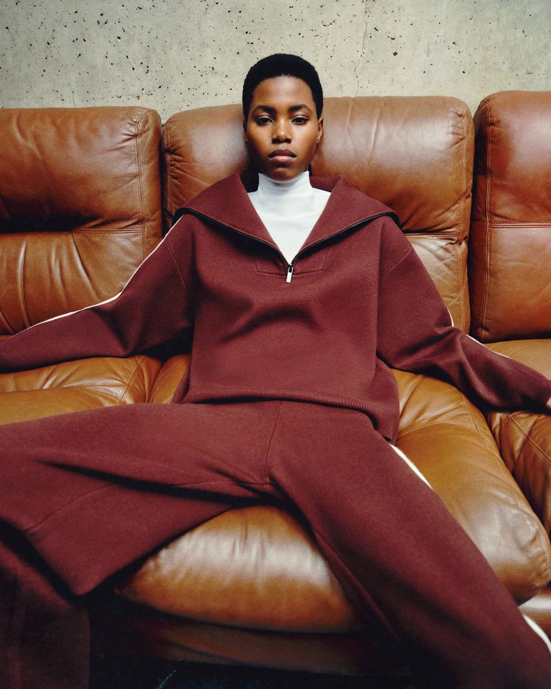 Young woman sits on a tan leather sofa, wearing a maroon tracksuit and white turtleneck, looking at the camera in an indoor setting.