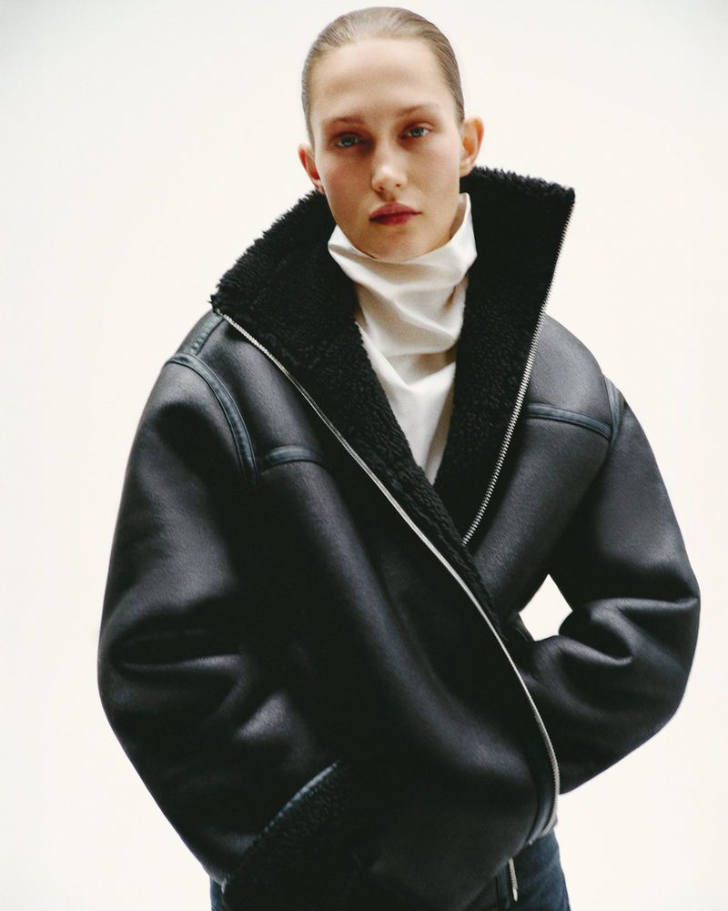 Fashion shot of a woman wearing a black bomber jacket with a white turtleneck, posing in a bright indoor photo studio setting.