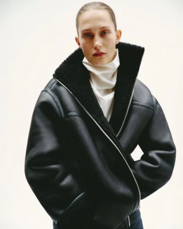 Fashion shot of a woman wearing a black bomber jacket with a white turtleneck, posing in a bright indoor photo studio setting.