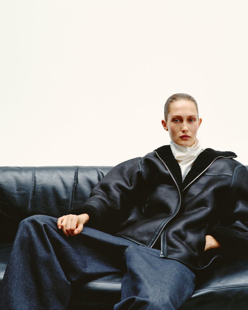 Adult female model poses on a black leather couch wearing wide-leg jeans and a black shearling jacket against a white background.