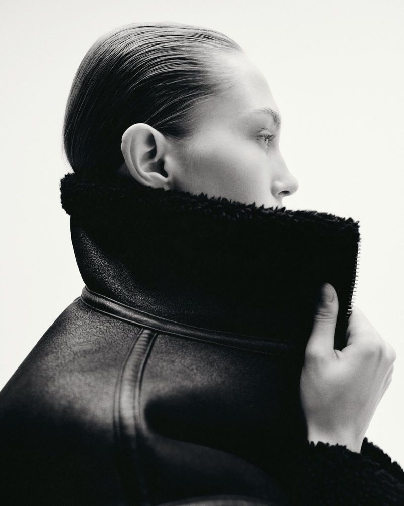 Monochrome portrait of a woman with slicked-back hair wearing a textured black coat with fur trim against a clean white background.
