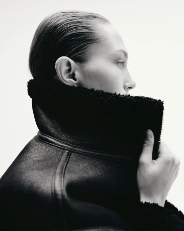 Monochrome portrait of a woman with slicked-back hair wearing a textured black coat with fur trim against a clean white background.