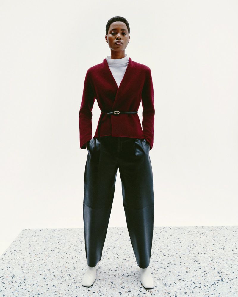 Fashion shot: A single woman in a wine-colored cardigan with a belt, black balloon pants, and white square-toed boots stands against a white backdrop.