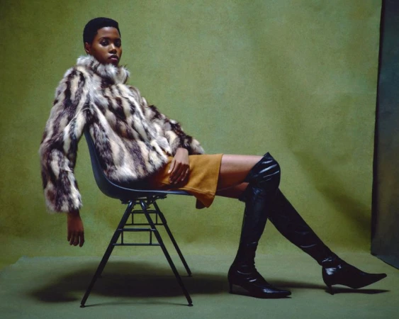 Model Victoria Fawole seated in a black chair wearing a patterned fur jacket, mustard skirt, and thigh-high boots against an olive backdrop.