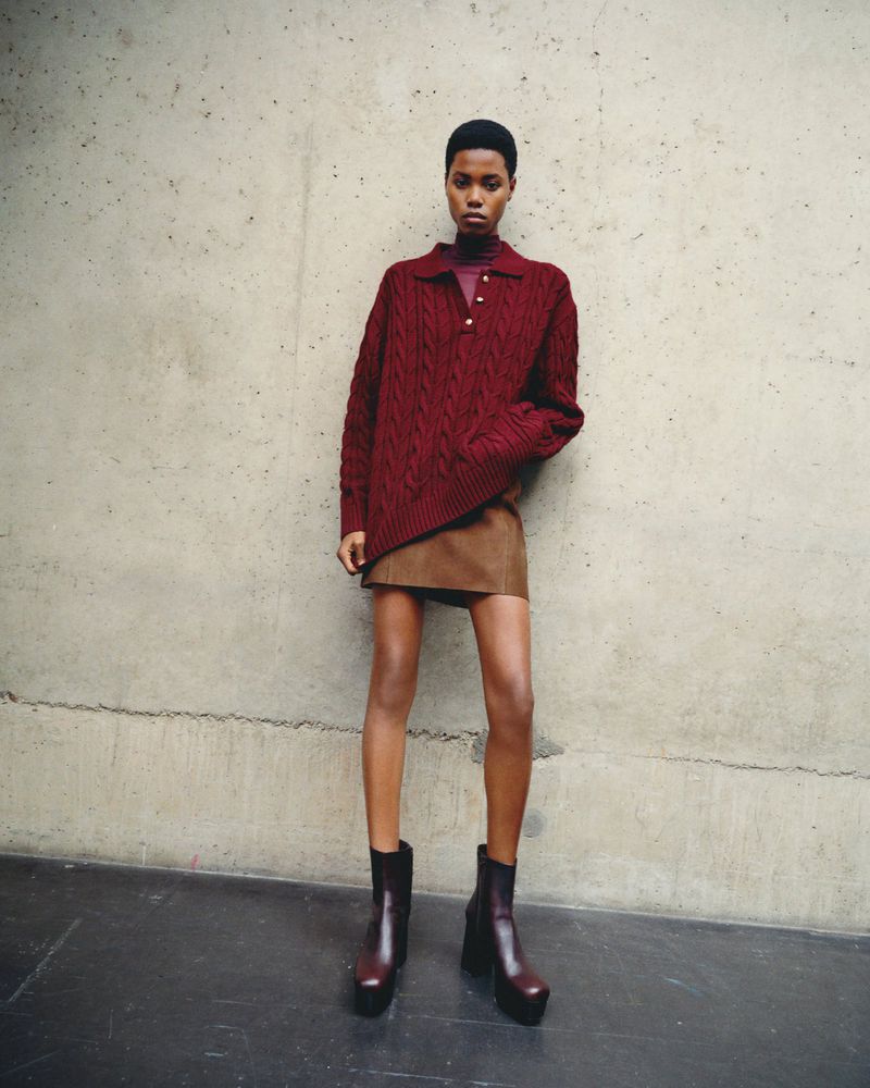 A full shot of a Black female model posing in a maroon cable-knit sweater, short brown skirt and maroon platform boots in a studio.