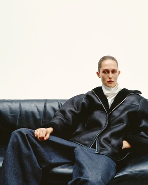 Model Vika Evseeva in an oversized black shearling jacket and wide-leg denim jeans seated on a black leather sofa against a white backdrop.