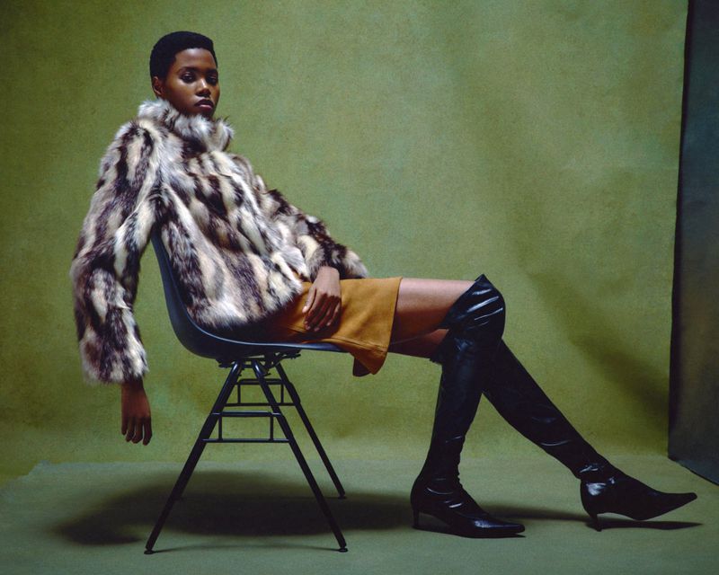 Model Victoria Fawole seated in a black chair wearing a patterned fur jacket, mustard skirt, and thigh-high boots against an olive backdrop.