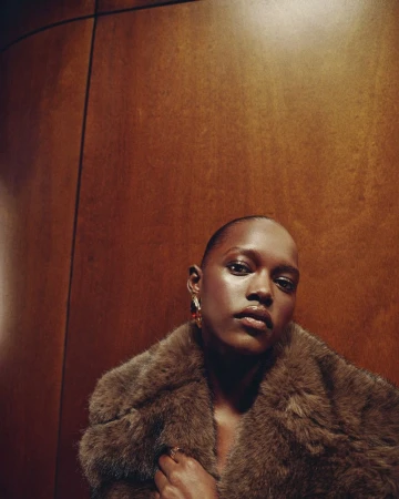 Stylish portrait of an adult female with short hair wearing a fur coat in front of a brown wood panel backdrop, looking directly at the camera.