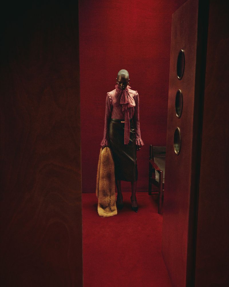 A woman in a magenta blouse and black skirt stands in a red corridor holding a fur coat, near a chair and a wall with oval cutouts.