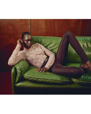 Fashion shot of a Black female model in textured top and leggings, reclining on a green leather couch against a dark wood-paneled wall.