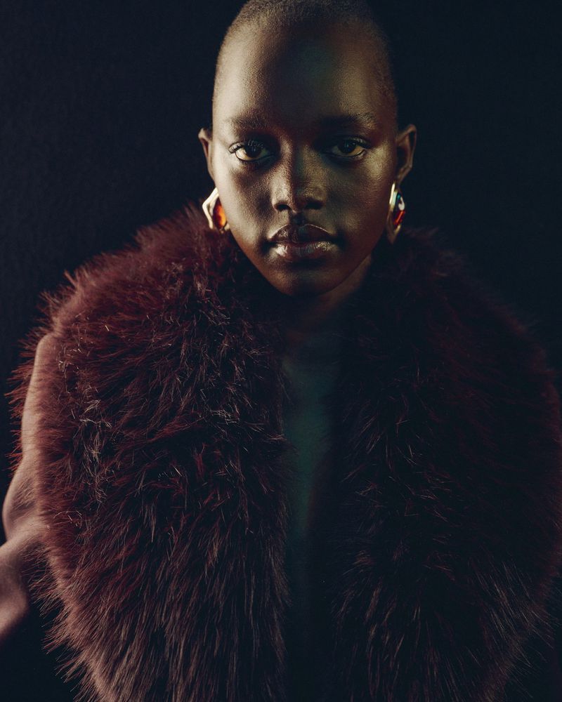 Fashionable portrait of a Black woman with short hair, wearing large geometric earrings and a feathery maroon coat, against dark backdrop.